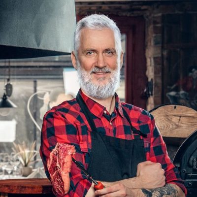 elderly meat shop worker with apron and uncooked meat e1674721188225